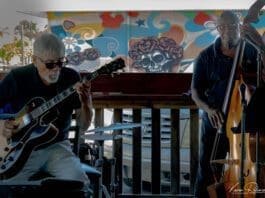 Two men playing instruments in front of a colorful wall.